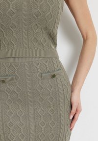 Oliivivihreä neulemekko, jossa on kabelineulekuvio, ribbireunus, kaksi taskua kontrastivärein, sekä kultasävytteiset nappikoristeet.