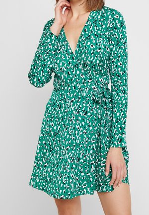 Femme portant une robe portefeuille verte à manches longues avec un motif léopard blanc et noir et une ceinture nouée à la taille, debout avec une main près du visage.