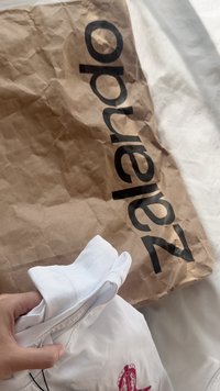 Camisa branca dobrada com um logótipo vermelho, segurada acima de um saco de papel castanho amassado com um grande texto preto a dizer "Zalando". Superfície de fundo suave.