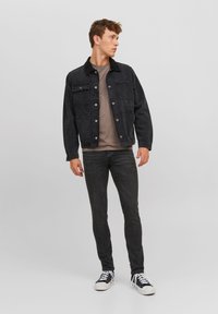 Jachetă din denim neagră cu două buzunare pe piept și închidere cu nasturi, purtată deasupra unei tricouri gri. Asociată cu blugi negri și adidași negri.