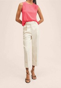 Ärmelloses, leuchtend pinkes Top mit drapiertem Design, kombiniert mit maßgeschneiderten, cremefarbenen Hosen mit umgeschlagenem Saum und zwei vorderen Falten. Schwarze Riemchensandalen.