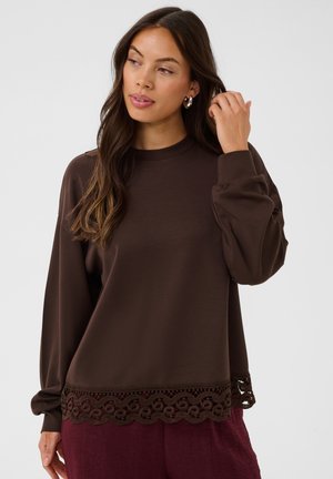 Femme aux longs cheveux bruns portant un haut marron ample avec une finition en dentelle à l'ourlet, un pantalon bordeaux et des créoles, touchant ses cheveux.