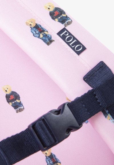 Sac à dos en tissu rose avec des graphiques d'ours bleu, une bandoulière marine et une étiquette "POLO" cousue sur le côté. Texture douce et design durable.