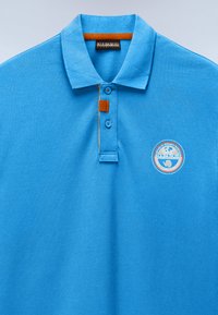 Μπλε πόλο πουκάμισο με δύο κουμπιά, μια μικρή πορτοκαλί ετικέτα και ένα κυκλικό λογότυπο Έρευνας Αρκτικής στο αριστερό στήθος.