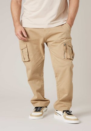 Homme portant un pantalon cargo beige à multiples poches et des baskets beige et blanc avec des lacets noirs, debout les mains dans les poches.
