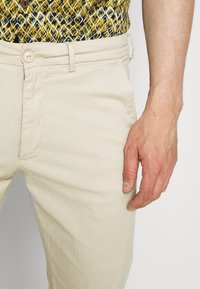 Beige bomuldsbukser med en glat tekstur, der har en knaplukning. En hånd er placeret på taljen, hvilket fremhæver pasformen og stoffet.