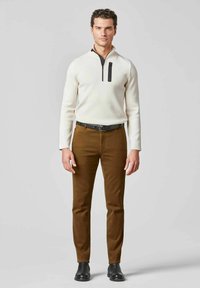 Weißer Reißverschluss-Pullover mit schwarzen Reißverschlussdetails, kombiniert mit braunen geraden Hosen und schwarzen Ankle Boots. Modell steht vor einem grauen Hintergrund.