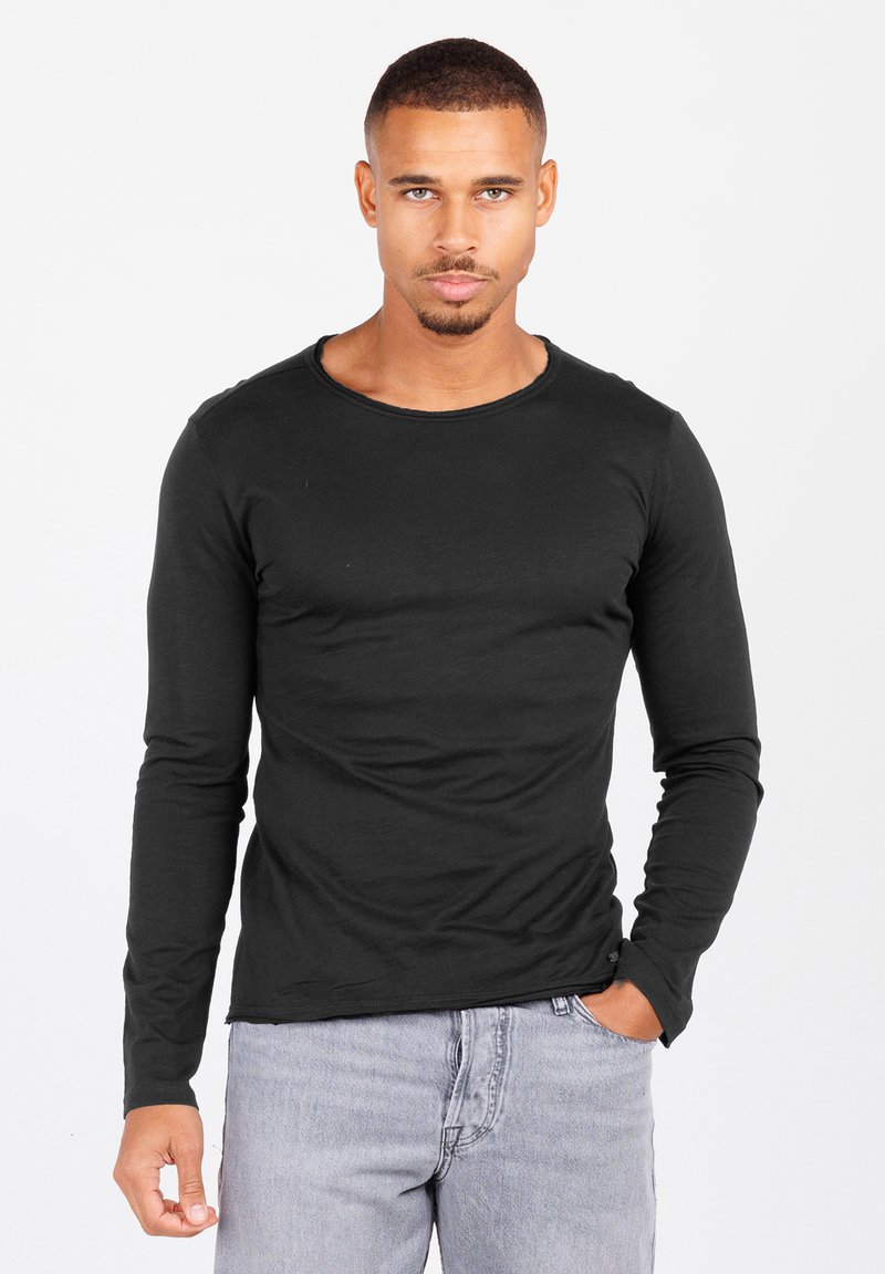 Schwarzes Langarm-T-Shirt aus weicher Baumwolle mit Rundhalsausschnitt und leicht ausgefranstem Saum. Trägt man zu hellgrauen Jeans.