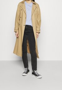 Trench-coat beige à double boutonnage, boutons noirs, manches retroussées, associé à un t-shirt bleu clair, un jean noir et des baskets noires.