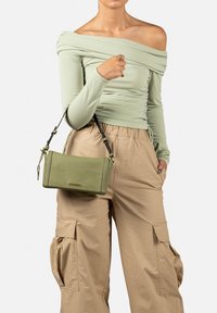 Borsa a tracolla in pelle verde con texture, dotata di una tracolla rimovibile e di forma rettangolare, abbinata a pantaloni cargo beige e una maglietta verde chiaro.