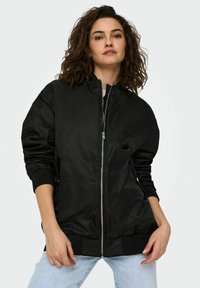 Veste aviateur noire avec fermeture éclair, poignets côtelés et coupe décontractée. Fabriquée dans un matériau lisse et léger. Comprend des poches latérales.