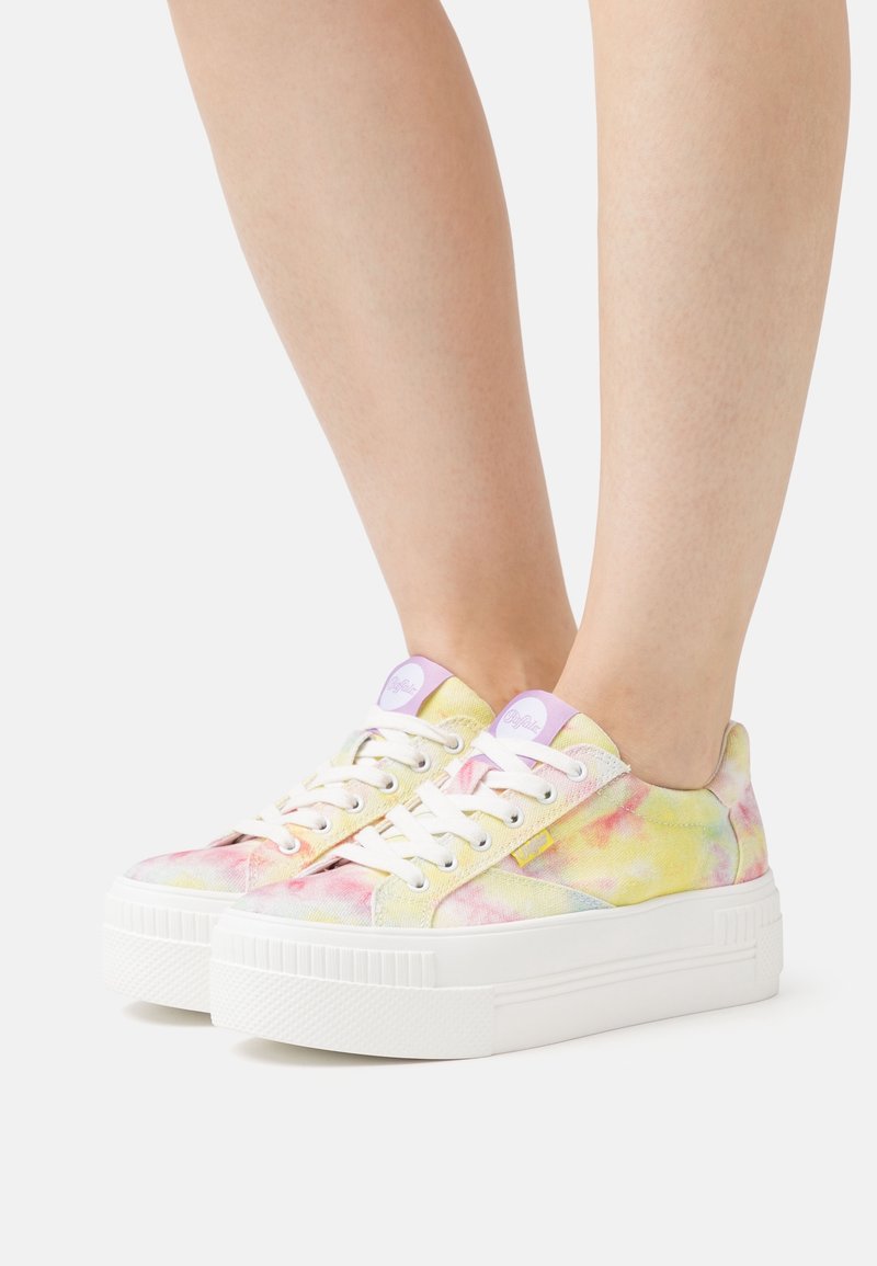 Pieds portant des baskets compensées tie-dye pastel avec des lacets blancs et des semelles blanches épaisses, présentés sur un fond blanc uni.