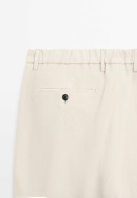 Pantalon beige clair avec une taille élastique, des passants de ceinture et une poche arrière passepoilée fermée par un bouton noir.