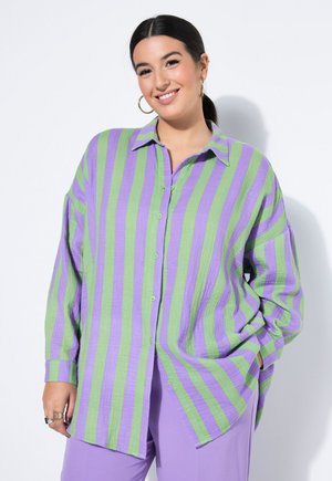 Lächelnde Frau mit dunklen Haaren im Pferdeschwanz trägt Creolen, ein locker sitzendes Hemd mit grünen und lila vertikalen Streifen und lila Hosen.