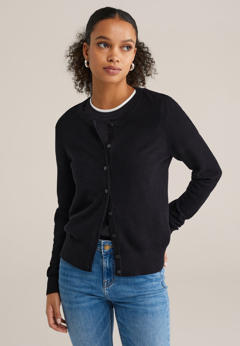 Crni pleteni kardigan s teksturiranim uzorkom, zaobljenim izrezom i kopčanjem na gumbe. Nošen preko crne majice i uparen s plavim trapericama.