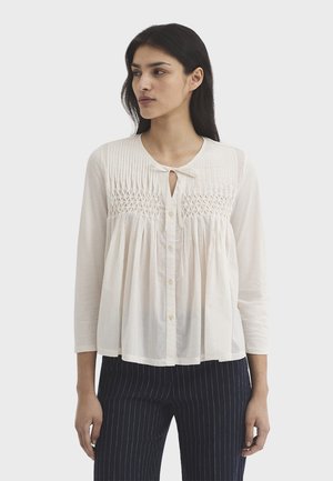 Mujer vistiendo una blusa crema de manga larga con detalles plisados y fruncidos, combinada con pantalones negros de rayas finas, de pie frente a un fondo liso.