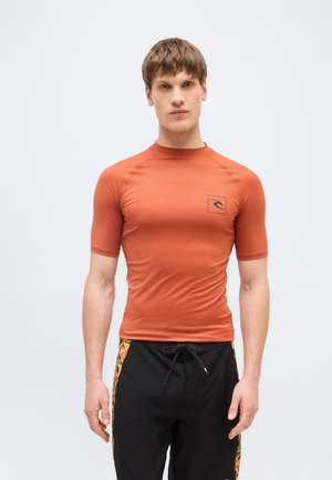 Jonge man die een strakke, roestkleurige rashguard met korte mouwen draagt, voorzien van een klein logo op de borst en zwarte korte broek met patroon op de zijkanten.