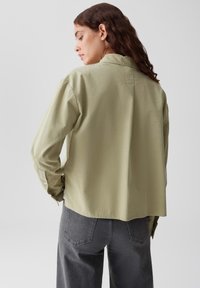 Eine Frau mit langen braunen Haaren, die ein lockeres, hellgrünes Langarmshirt und dunkle graue Jeans trägt, ist von hinten dargestellt und schaut zur Seite.