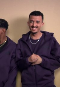 Two men wearing purple hoodies stand against a beige wall, one smiling and clasping his hands while the other looks down.