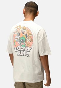 Bílý bavlněný tričko s barevným grafickým potiskem Looney Tunes na zadní straně, zobrazující postavy na jemném oranžovém kruhovém pozadí.