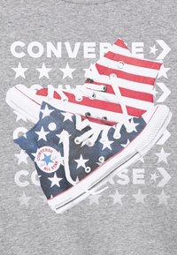 T-shirt grafico con due sneaker illustrate. Uno ha righe rosse e bianche, l'altro è blu con stelle bianche, su uno sfondo grigio.