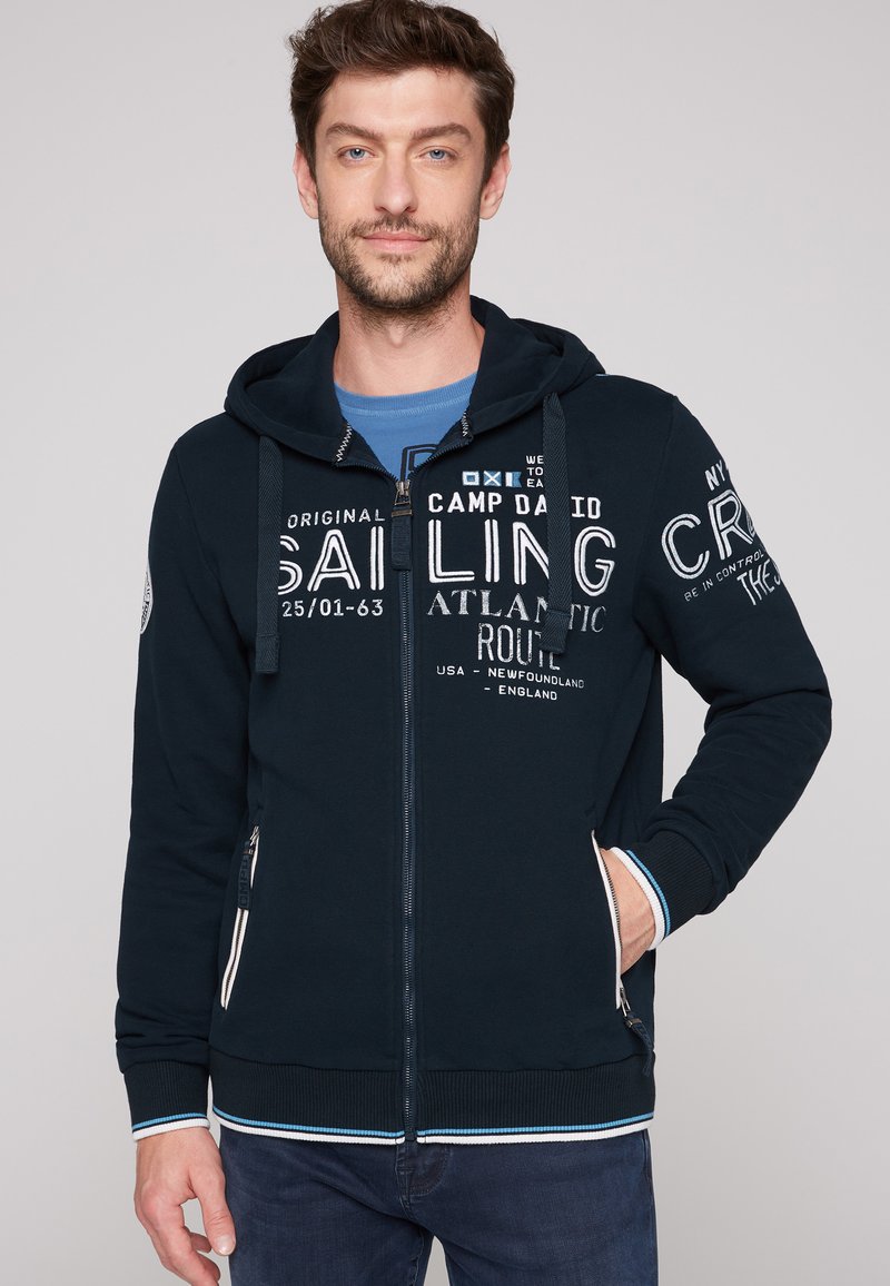 Felpa con zip di colore blu navy con testi bianchi, cappuccio con cordino e polsini a righe. Presenta due tasche laterali con zip e una toppa con logo sulla manica.