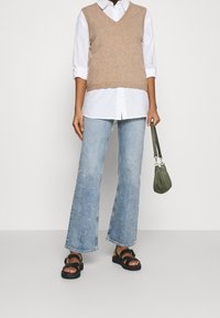 Chaleco de punto beige sobre una camisa blanca de botones, combinado con jeans acampanados de color azul claro y sandalias negras. Los accesorios incluyen un bolso verde.