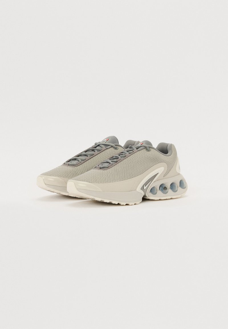 Zapatillas deportivas de color gris claro con parte superior de malla texturizada, punta redonda y suelas con amortiguación de aire transparente. Presentan cordones grises y un acento de logo.