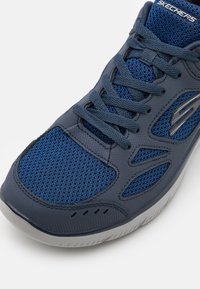 Sapatilha atlética Skechers com parte superior em malha azul-marinho, design texturizado, realces em cinzento e uma sola de borracha branca com padrão de aderência.