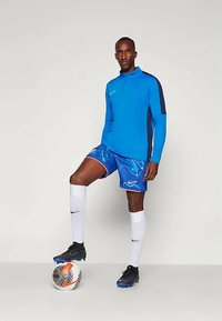 Blå långärmad träningsöverdel med marinblå detaljer, ihop med blå mönstrade shorts. Vita knästrumpor och svarta fotbollsskor, med en fotboll.