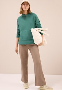 Mujer con una sudadera verde, pantalones de pata ancha beiges, gorra beige y zapatillas, sosteniendo una chaqueta de cremalleras acolchada color crema frente a un fondo liso.