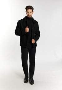 Schwarzer Mantel mit Kragen-Design, ausgestattet mit Druckknöpfen und seitlichen Taschen. Getragen über einem schwarzen Rollkragenpullover und kombiniert mit schwarzen Hosen und Schuhen.
