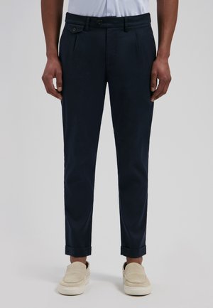 Man draagt donker marineblauwe op maat gemaakte broek met omgeslagen zoom, gecombineerd met beige instapschoenen op een effen lichtgrijze achtergrond.