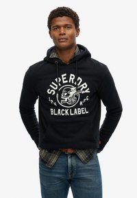 Sudadera negra con un gráfico frontal que presenta una calavera y el texto "SUPERDRY BLACK LABEL". Llevada sobre una camisa de cuadros y combinada con unos vaqueros azules.