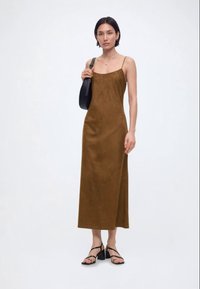 Γυναίκα που φοράει καφέ αμάνικο μίντι φόρεμα, μαύρα ψηλοτάκουνα πέδιλα με λουράκια και κρατά μια μαύρη τσάντα ώμου σε λευκό απλό φόντο.