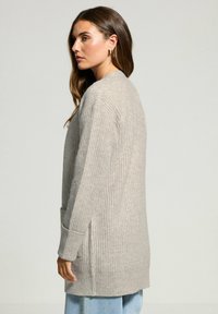 Cardigan lungo aperto in maglia a coste beige chiaro. Presenta tasche laterali e maniche a sbuffo. Texture morbida e vestibilità rilassata.