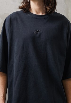 Personne portant un t-shirt noir Nike avec un petit logo Nike brodé centré sur la poitrine, sur un fond neutre.