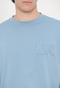Vaaleansininen puuvillapaita pyöreällä kaula-aukolla, jossa on teksturoitu brodeerattu "MK" -logo vasemmassa rinta-alueessa.