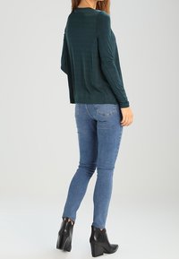 Langärmliges, tiefgrünes Oberteil mit glatter Textur, kombiniert mit blauen Skinny-Jeans und schwarzen, hohen Stiefeletten. Lässiges Outfit von hinten.