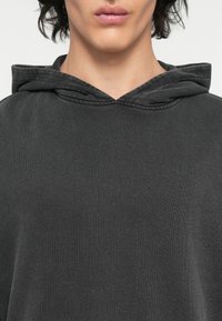 Sweat à capuche gris foncé en tissu texturé, avec un col large, une capuche à cordon et une coupe décontractée. Design simple sans motifs visibles.