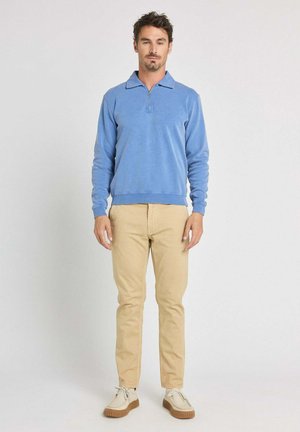 Homme debout, portant un pull léger bleu clair à manches longues avec fermeture éclair et un pantalon beige, sur un fond clair uni.