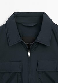Dark navy jacket featuring a fold-down collar, front zipper, and two chest pockets with flap detail. Smooth, padded texture.