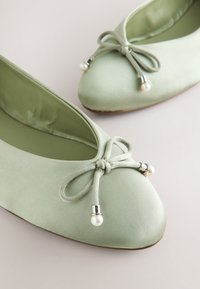 Ballerines en satin vert clair avec des nœuds ornés de perles et de perles argentées sur une surface neutre.