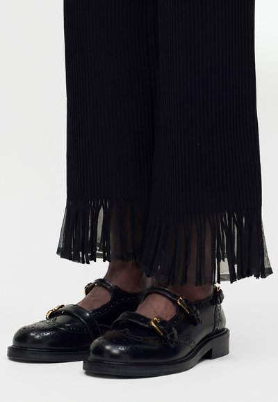 Μαύρα δερμάτινα brogues Mary Jane με χρυσές αγκράφες, συνδυασμένα με μαύρο πλισέ παντελόνι με διάφανα, κρόσσια στα τελειώματα.