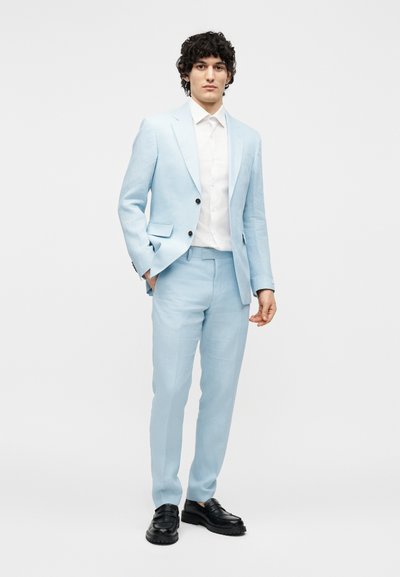 Νέος άνδρας που στέκεται, φοράει ανοιχτό μπλε κοστούμι με άσπρο πουκάμισο και μαύρα επίσημα παπούτσια σε απλό λευκό φόντο.