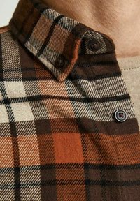 Camicia di flanella a quadri marrone e arancione con colletto a bottone, caratterizzata da texture a contrasto e un mix di modelli a scacchi.