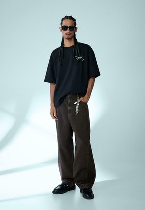 Uomo con capelli intrecciati e occhiali da sole, che indossa una maglietta nera oversize e pantaloni scuri larghi con un portachiavi agganciato alla tasca.