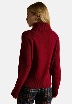 Roter gestrickter Pullover mit hohem Rippkragen, gerippten Bündchen und Saum, der Raglanärmel besitzt. Weiche Textur vor einem schlichten Hintergrund.