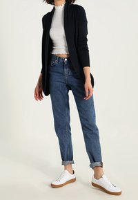 Schwarze offene Strickjacke, weißes, kurz geschnittenes, geripptes Top, blaue hochtaillierte Jeans und weiße Sneakers mit braunen Sohlen, lässig gestylt.