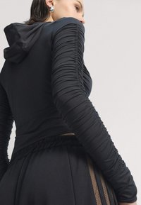 Black hooded top with ruched long sleeves, paired with black pleated pants featuring a sheer panel on the side. Smooth, stretchy fabric.